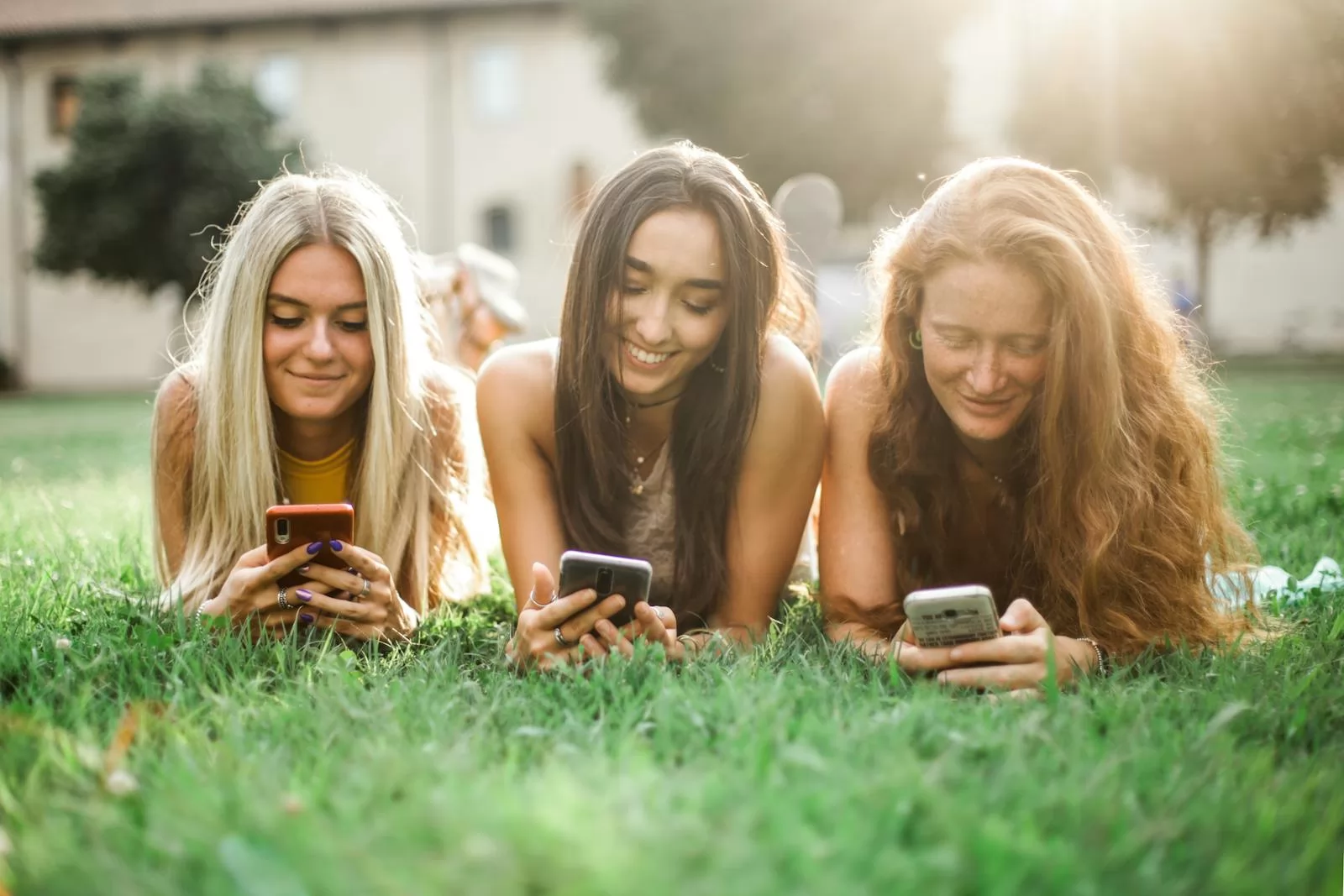 Three young women relaxing on grass using smartphones in a sunny park.