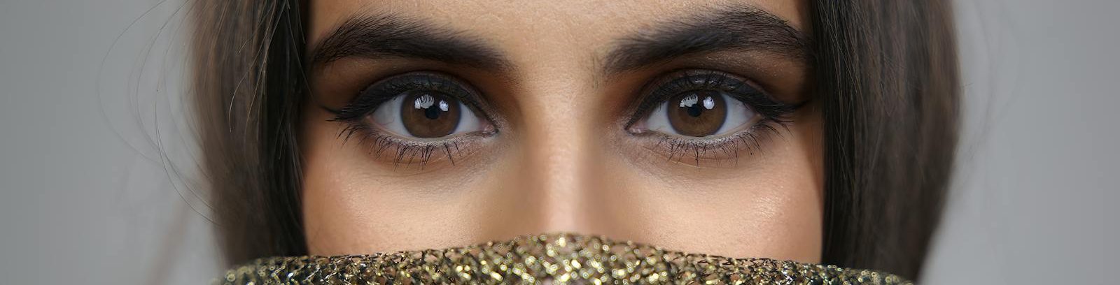 A close-up portrait highlighting a woman's expressive brown eyes and gold scarf in a soft studio setting.