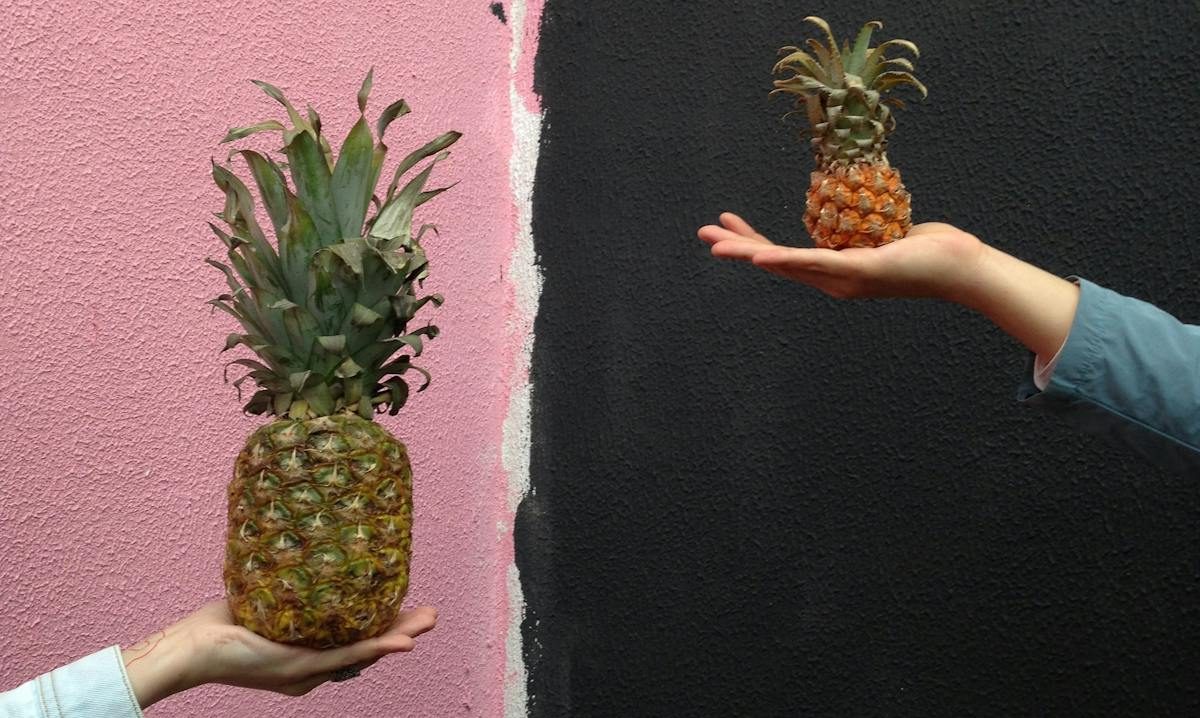 Two hands hold different sized pineapples against a colorful textured wall, showcasing a size contrast theme.